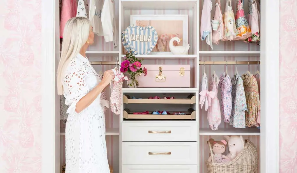 a woman holding a baby onesie looking at a custom baby closet with pink accents
