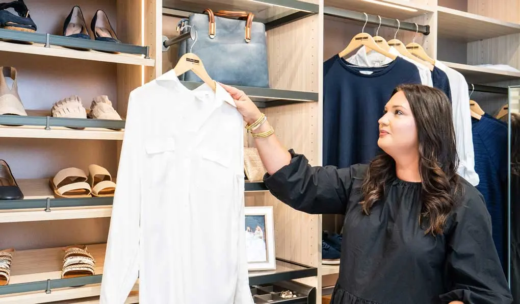 A woman pulling out white button up shirt in a light beige wood closet with custom shelving